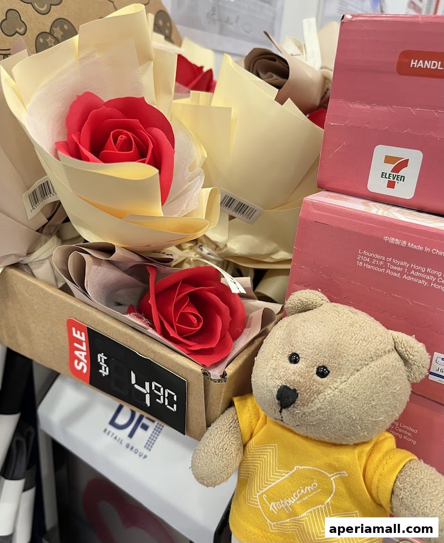 A stuffed bear wearing a yellow shirt stands next to bouquets of red roses and 7-Eleven branded boxes.