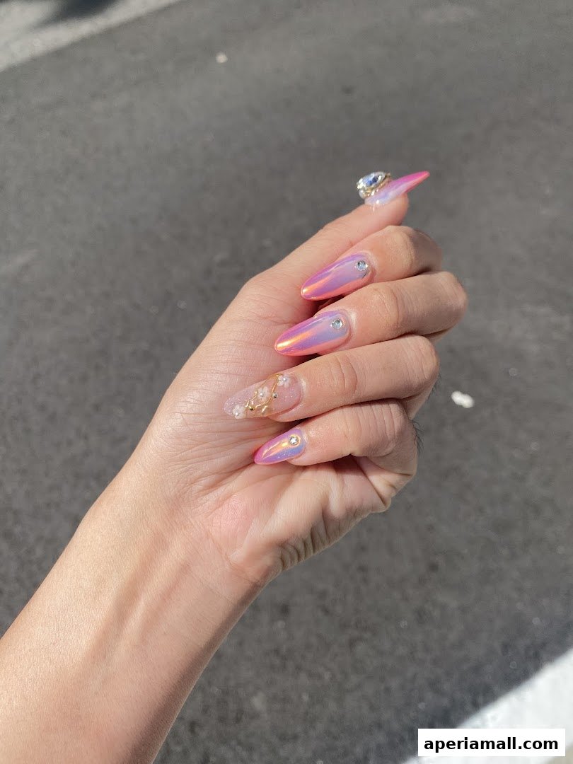 A hand with long, almond-shaped nails featuring iridescent pink polish and rhinestones.