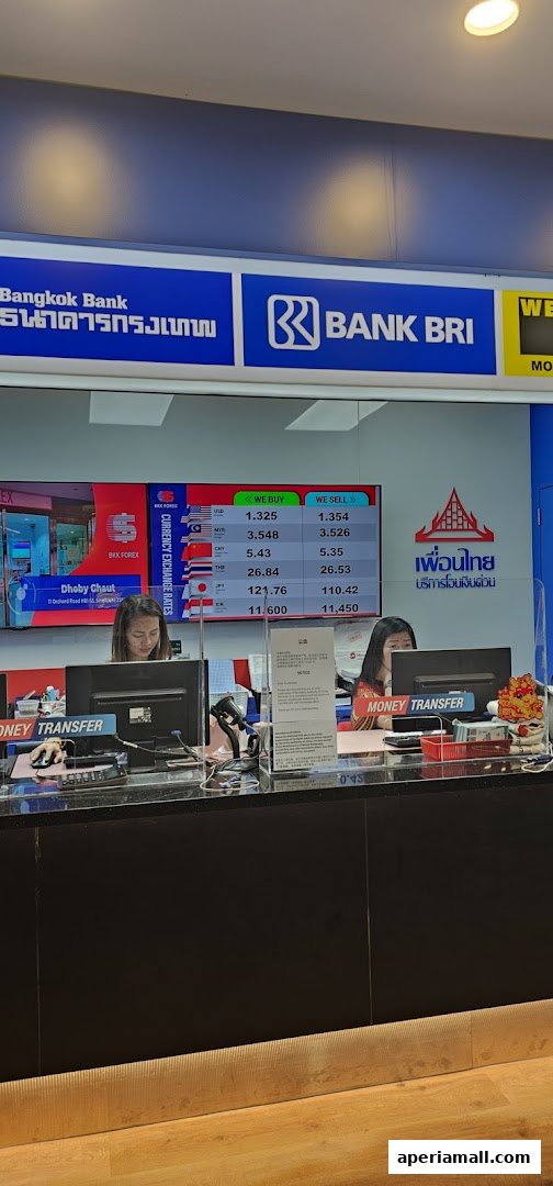 Two women work at counters with computers displaying currency exchange rates and bank logos.