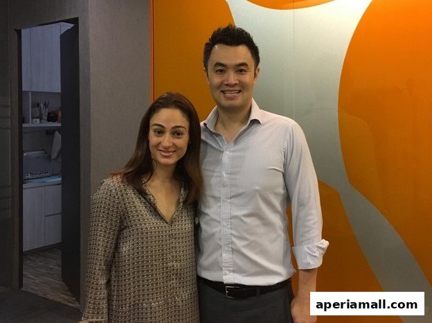 A man and a woman smiling in front of an orange and grey wall.