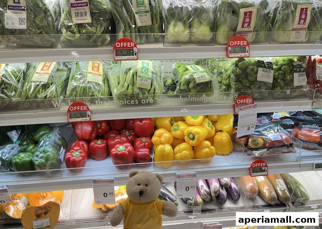 A display of fresh vegetables including leafy greens, bell peppers, and eggplants, with a teddy bear mascot.