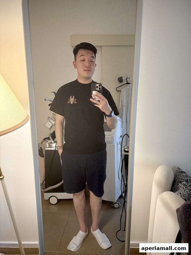 A man in a black t-shirt and shorts takes a mirror selfie, showing off his outfit.
