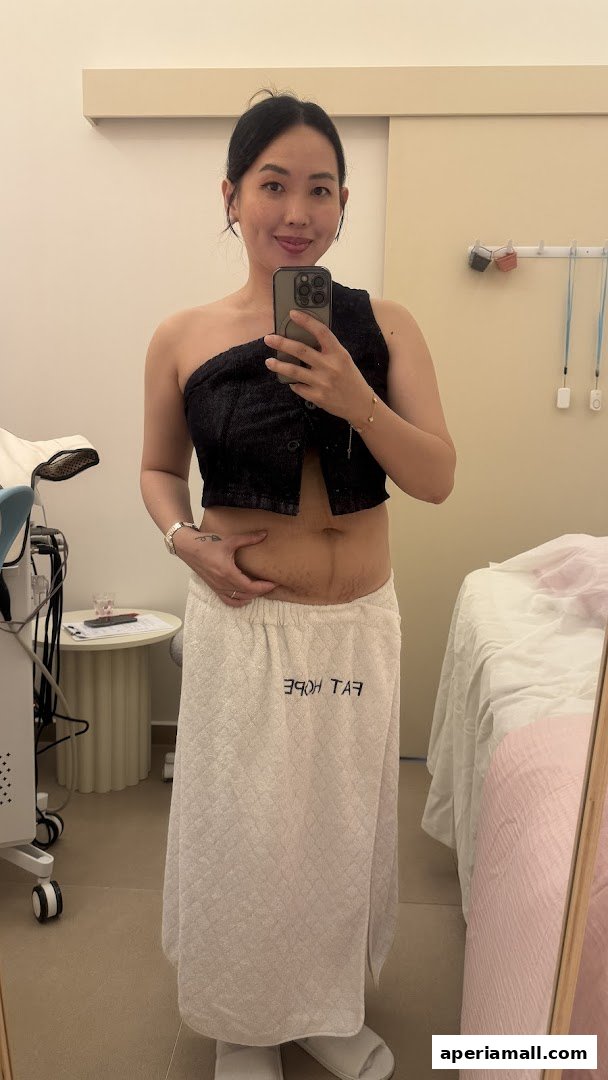 A woman in a black top and white towel poses for a mirror selfie in a treatment room.