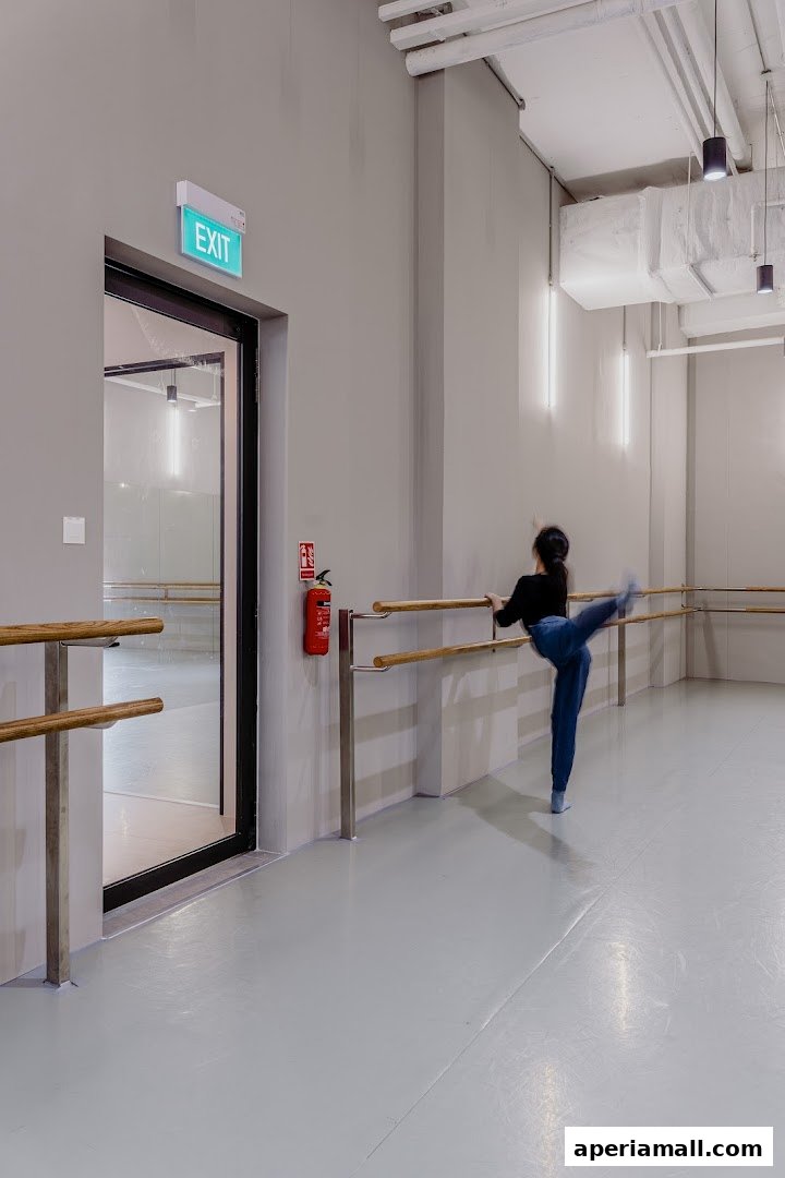 A dancer practices at a ballet barre in a spacious dance studio with an exit sign.