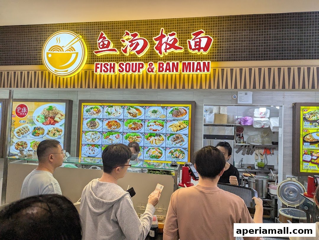 Customers ordering at a food stall with a large menu display featuring various noodle dishes.