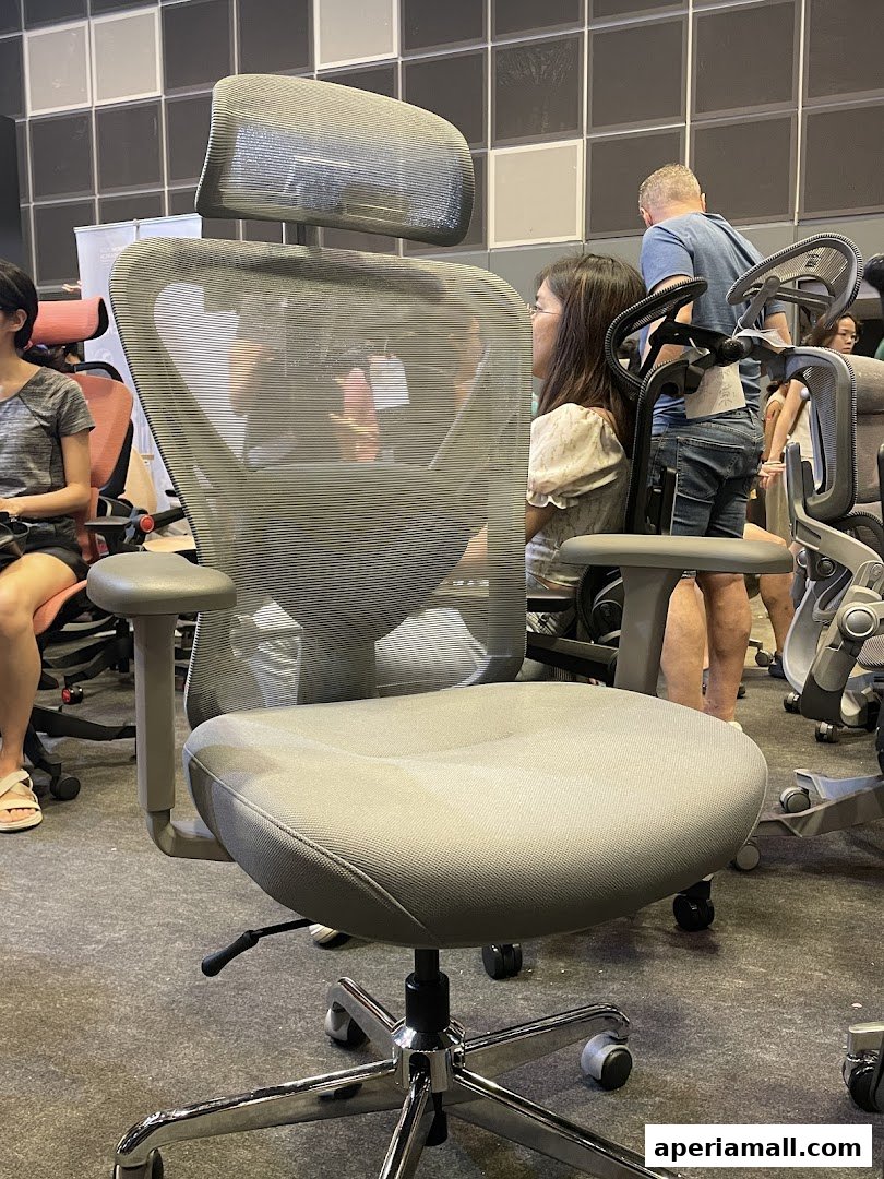A close-up of a grey ergonomic office chair with people browsing in the background.
