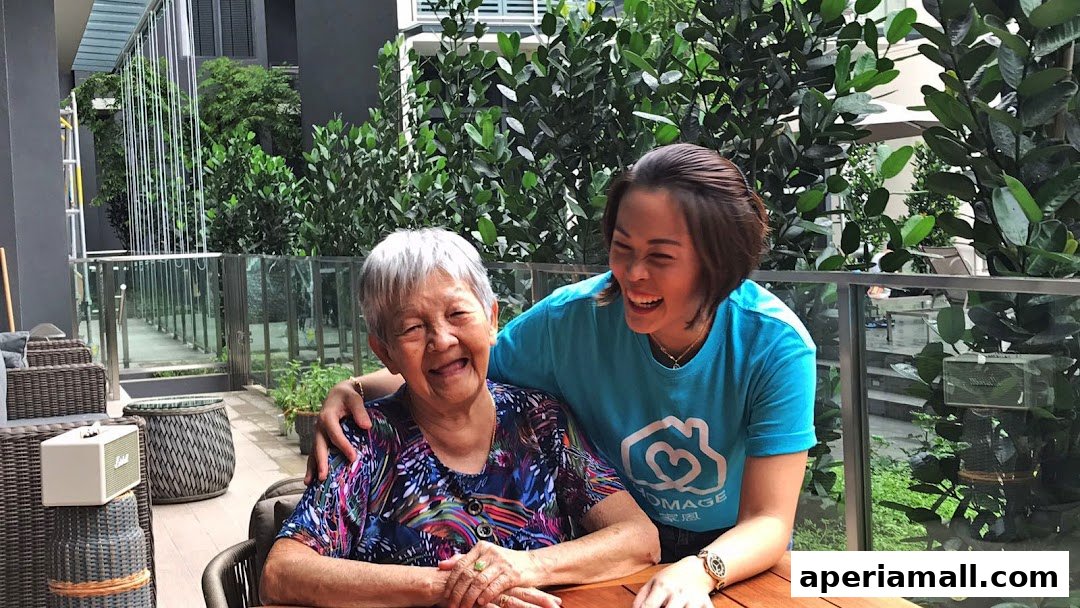 A younger woman in a blue t-shirt embraces an elderly woman, both smiling warmly.