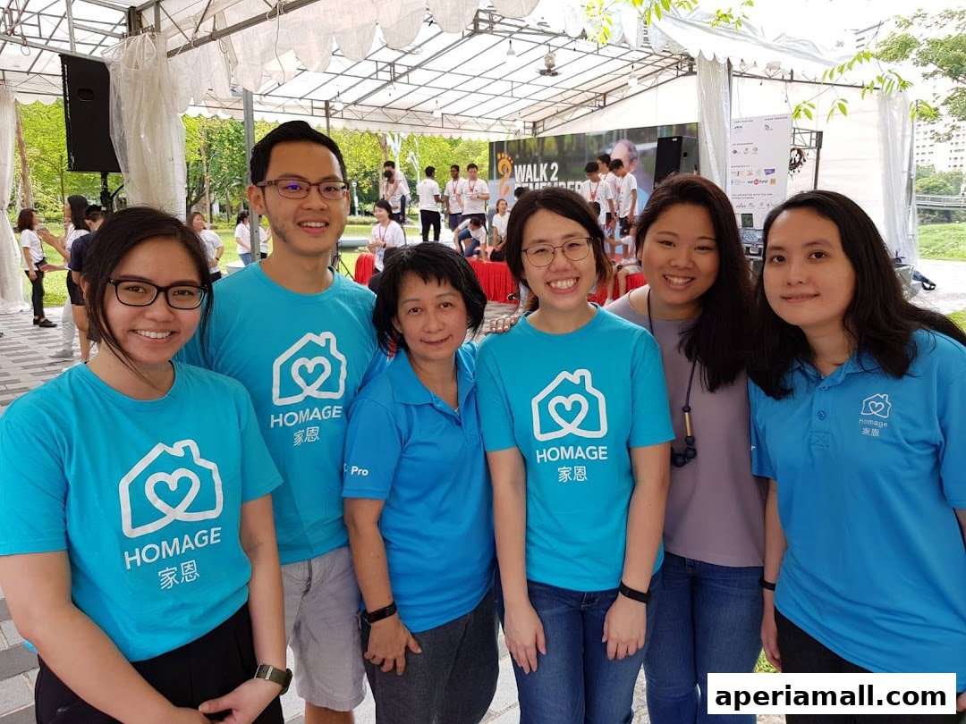 A group of smiling people wearing blue t-shirts with the Homage logo at an outdoor event.