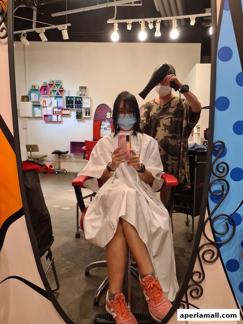 A person in a salon cape is getting their hair dried by a stylist reflected in a mirror.