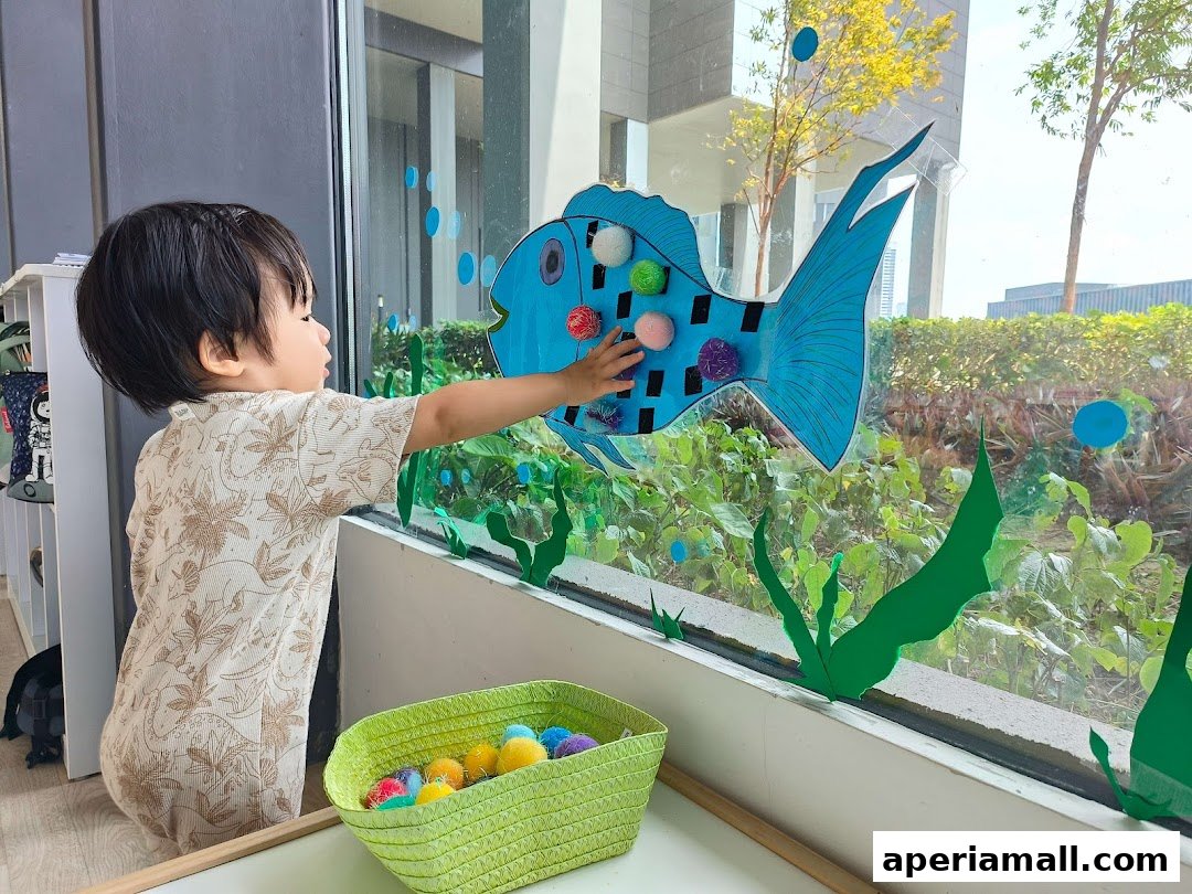 A young child interacts with a large blue fish decoration on a window, with a basket of colorful pom-poms nearby.