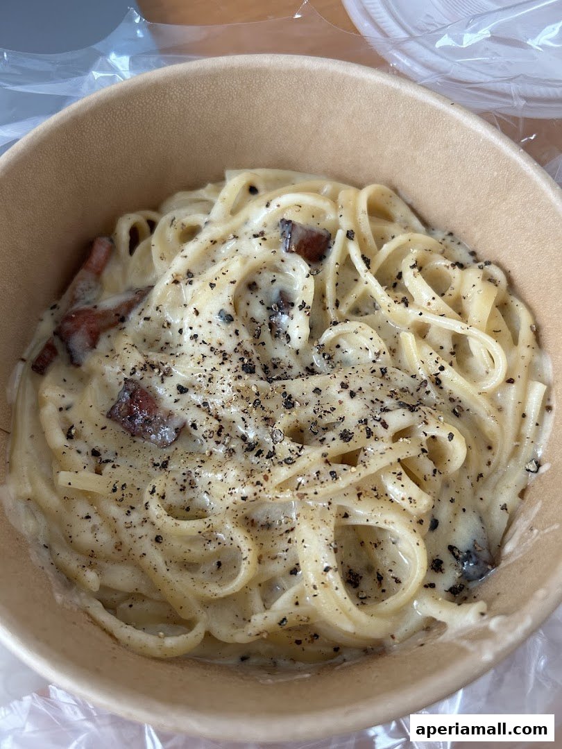 A close-up shot of creamy pasta with bacon and black pepper in a to-go container.