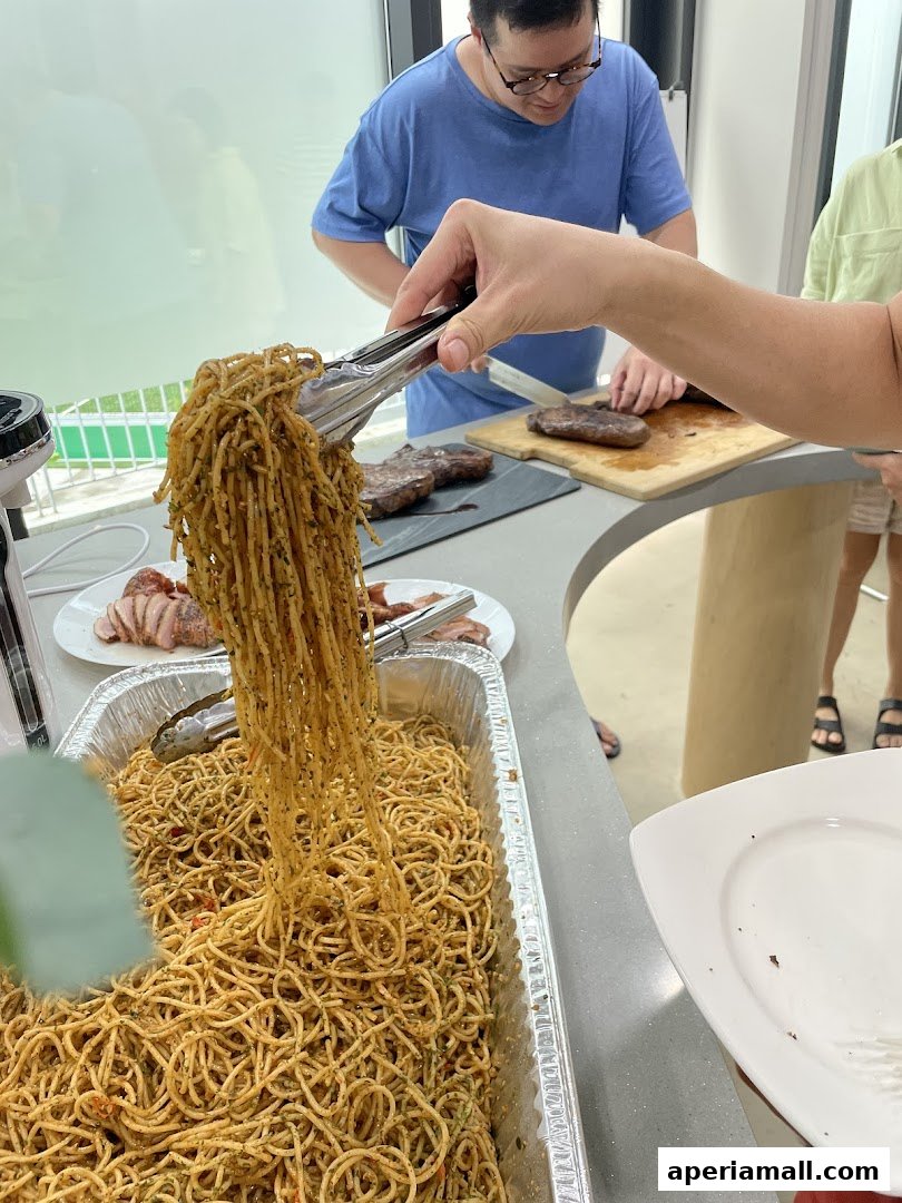 A person is serving spaghetti from a large tray at a gathering with steaks being prepared.