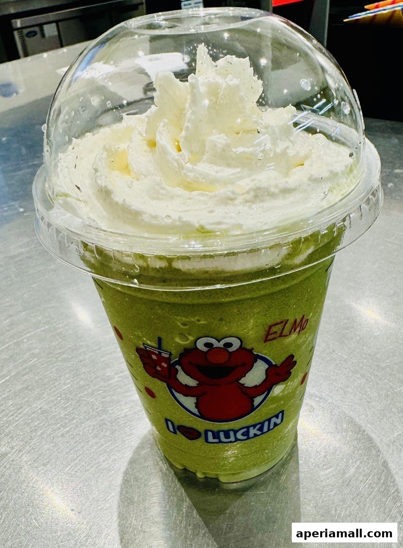 A close-up of a green beverage topped with whipped cream in a clear plastic cup with Elmo branding.