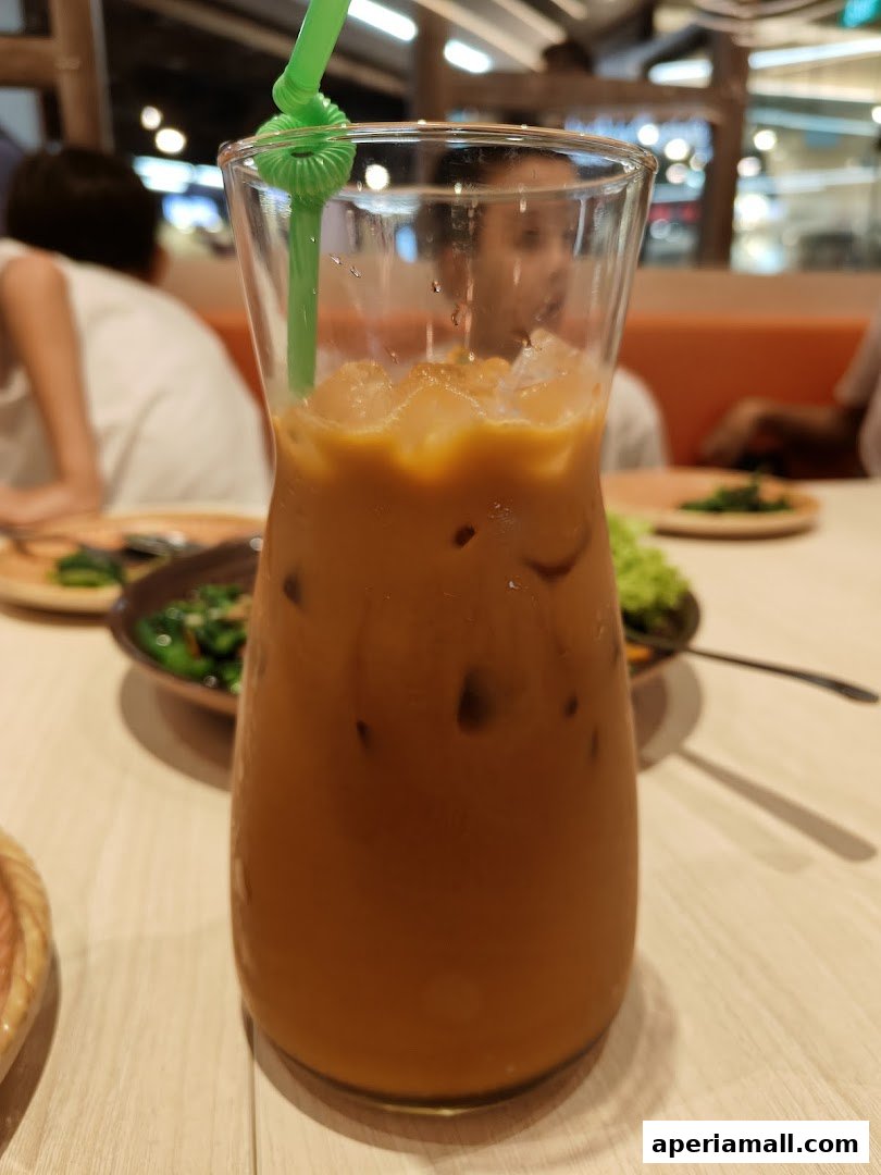 A tall glass of iced Thai milk tea with a green straw and ice cubes.