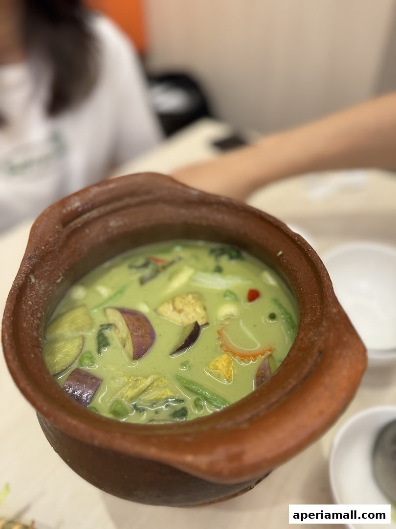 A close-up of a clay pot filled with green curry, featuring vegetables and tofu.