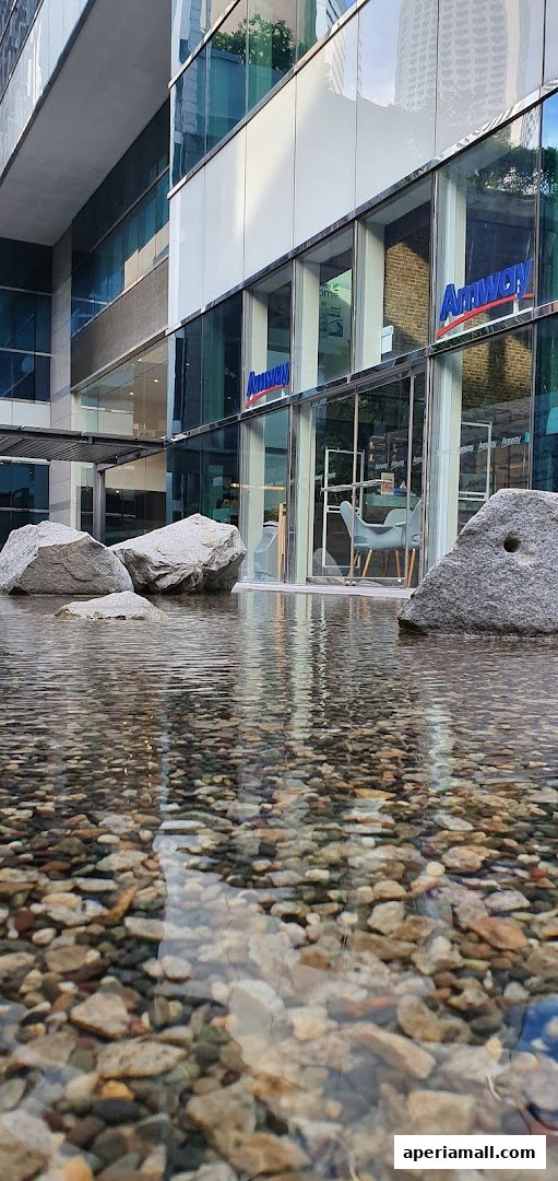 The exterior of an Amway shop at Aperia Mall, featuring large windows and a water feature with rocks.