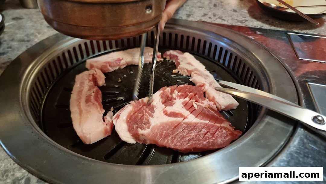 Raw pork belly being grilled on a Korean BBQ grill with tongs and scissors.