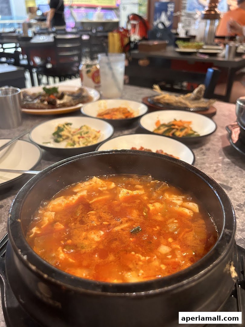 A steaming bowl of Korean tofu stew with various side dishes and a fried fish.