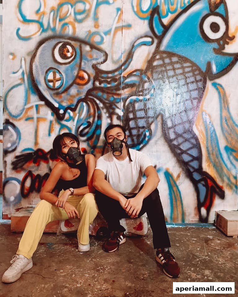 Two people wearing masks sit in front of a graffiti-covered wall with cartoonish fish and octopus art.