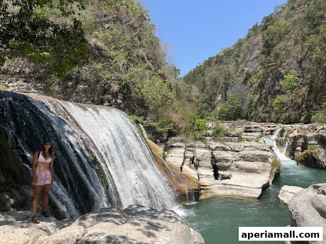 A woman stands by a cascading waterfall in a lush, rocky canyon.