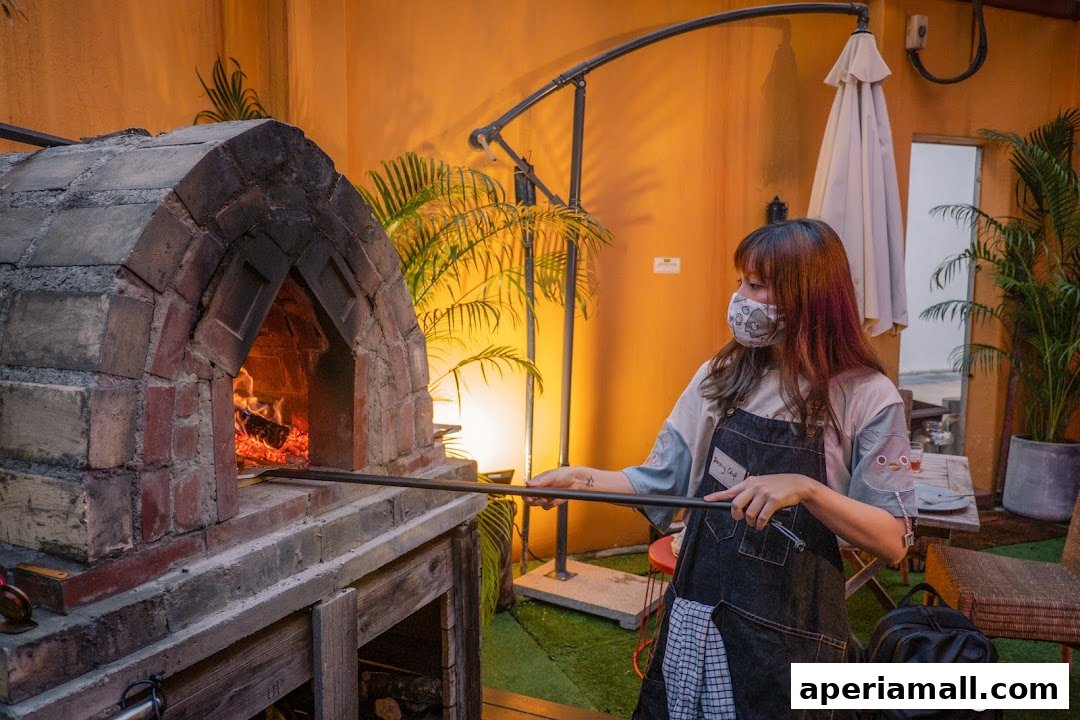 A woman in an apron uses a peel to tend to a wood-fired oven.