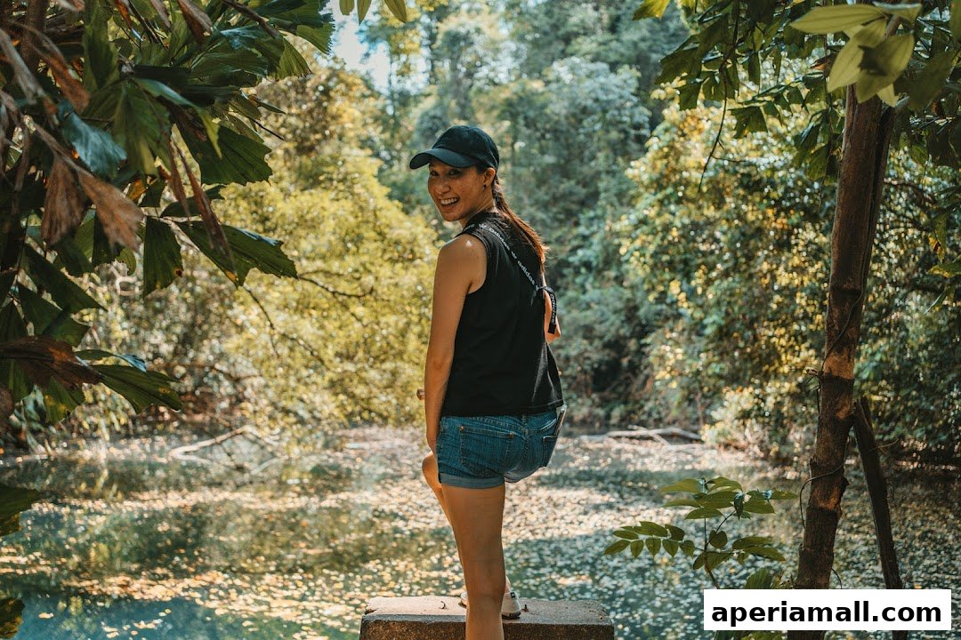 A woman in a black tank top and denim shorts smiles at the camera in a lush, green forest setting.
