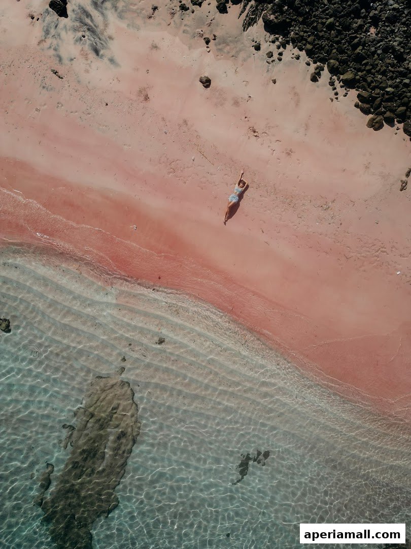Aerial view of a person relaxing on a pink sand beach next to clear turquoise water.