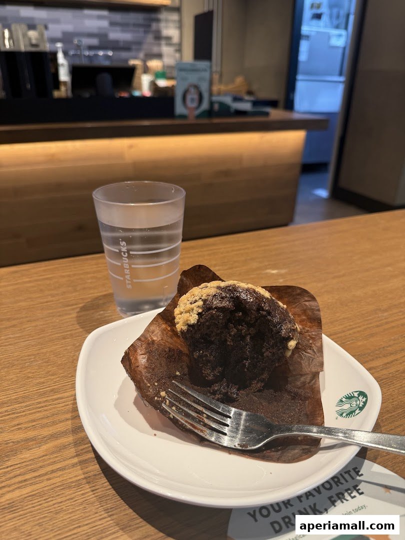 A chocolate muffin with a fork on a plate next to a glass of water at Starbucks.