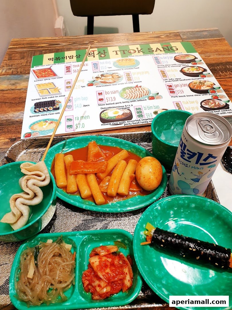 A table laden with Korean dishes, including tteokbokki, kimbap, and kimchi, with a menu and drink.