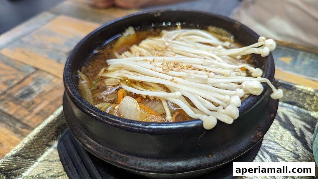 A close-up of a steaming bowl of Korean stew with enoki mushrooms and vegetables.