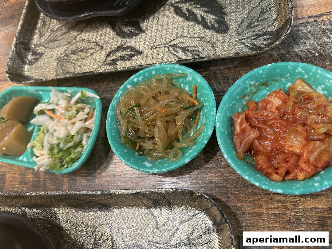 Three small bowls of Korean side dishes, including kimchi, japchae, and a salad, are arranged on a wooden table.