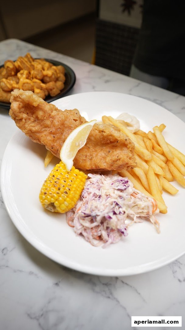 A plate of fish and chips with coleslaw, corn on the cob, and a lemon wedge.