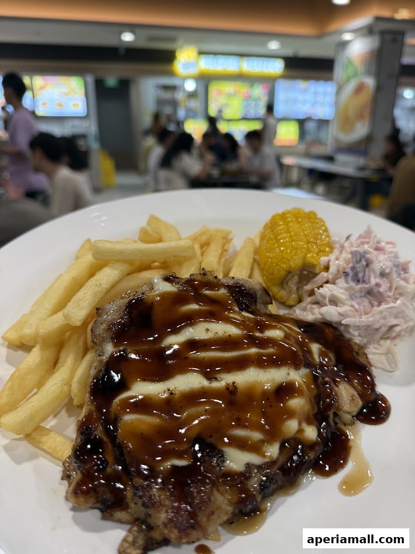 A plate of grilled chicken with fries, corn, and coleslaw, with a blurred food court background.