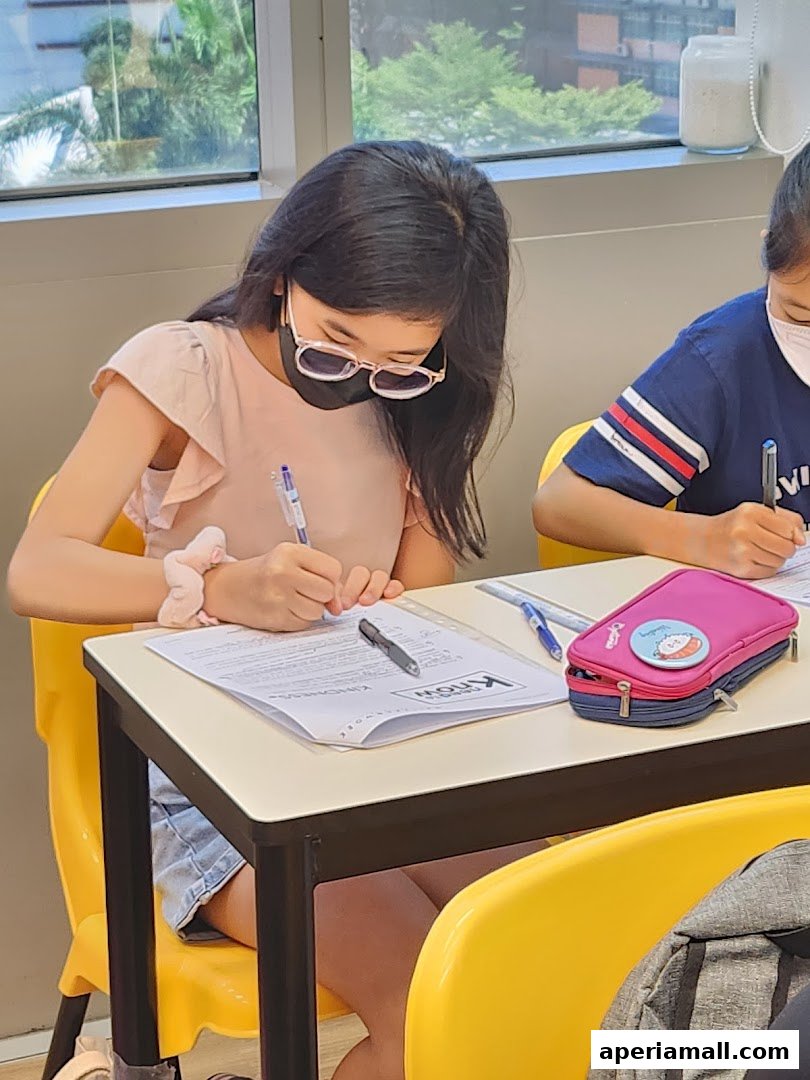 Students wearing masks focus on writing at desks in a bright classroom setting.